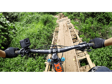 Vue à la première personne d'un cycliste sur un pont en bois au-dessus d'un ruisseau. La végétation verte entoure le pont.