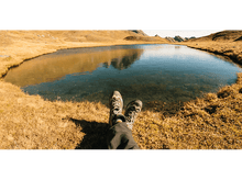 Charger l&#39;image dans la galerie, Jambes d&#39;une personne avec des chaussures de randonnée se reposant près d&#39;un lac, herbe sèche, montagnes en arrière-plan.
