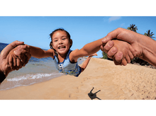 Charger l&#39;image dans la galerie, Un enfant est tenu par des mains et soulevé en l&#39;air sur une plage avec un ciel bleu.
