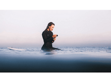 Charger l'image dans la galerie, Une femme en combinaison assise sur une planche de surf, regardant son téléphone dans l'océan.
