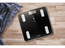 Charger l&#39;image dans la galerie, Une balance intelligente noire sur un sol en bois, avec un tissu gris en arrière-plan.
