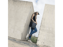 Charger l&#39;image dans la galerie, Une personne en casquette et jean s&#39;appuie contre un mur en béton. Elle porte un sac noir.
