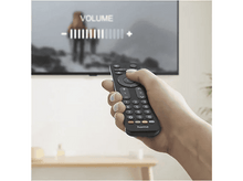 Charger l&#39;image dans la galerie, Une main tient une télécommande noire pour régler le volume de la télévision. L&#39;écran affiche une scène extérieure floue. Barre de volume visible.
