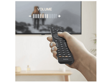 Charger l&#39;image dans la galerie, Main tenant une télécommande noire réglant le volume sur un téléviseur. Le téléviseur montre une scène floue de personnes.
