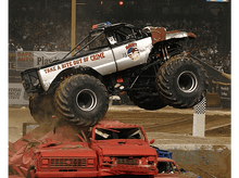 Charger l&#39;image dans la galerie, Monster truck sautant par-dessus des voitures écrasées lors d&#39;un événement de stade, avec une foule en arrière-plan.
