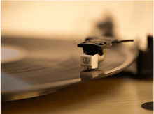 Charger l&#39;image dans la galerie, Gros plan d&#39;une platine vinyle avec bras noir et cellule blanche jouant un disque vinyle sur une surface en bois.
