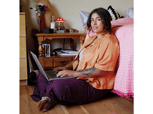 Charger l&#39;image dans la galerie, Une femme assise par terre utilise un ordinateur portable. Un lit et une table de chevet sont en arrière-plan.
