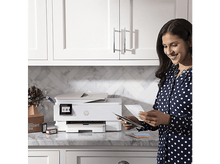 Charger l&#39;image dans la galerie, Femme en chemisier à pois regardant des papiers imprimés devant une imprimante et des cartouches d&#39;encre.
