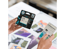 Charger l&#39;image dans la galerie, Des mains tiennent une boîte de cartouches d&#39;encre HP et un téléphone. Le téléphone affiche l&#39;application HP Smart.
