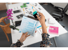 Charger l&#39;image dans la galerie, Mains tenant une cartouche d&#39;encre HP et une enveloppe imprimée sur une table blanche avec des documents.
