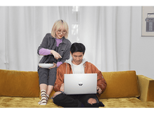 Charger l&#39;image dans la galerie, Un couple sur un canapé jaune regardant un ordinateur portable, l&#39;un pointant vers une assiette de nourriture.
