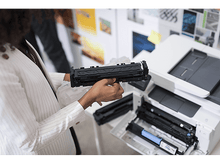 Charger l&#39;image dans la galerie, Une personne tient une cartouche de toner noire près d&#39;une imprimante. Le compartiment à toner de l&#39;imprimante est ouvert.
