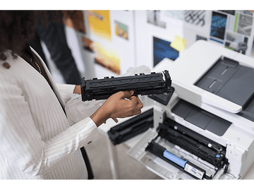 Une personne tient une cartouche de toner noire près d'une imprimante. Le compartiment à toner de l'imprimante est ouvert.