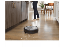 Charger l&#39;image dans la galerie, Un aspirateur robot sur un plancher en bois, avec une personne en arrière-plan près des meubles de cuisine.
