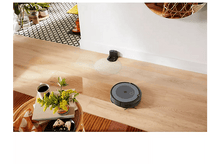 Charger l&#39;image dans la galerie, Aspirateur robot sur un sol en bois, près d&#39;une base de chargement et d&#39;une table avec des objets.
