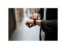 Charger l&#39;image dans la galerie, La main d&#39;une personne ajustant une montre de sport noire avec un bracelet noir au poignet, en extérieur.
