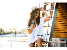 Charger l&#39;image dans la galerie, Une femme en robe blanche tient un téléphone près de l&#39;eau et d&#39;un bateau.
