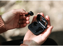 Charger l&#39;image dans la galerie, Mains tenant des écouteurs sans fil noirs et leur étui de chargement.
