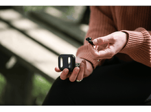 Charger l&#39;image dans la galerie, Personne tenant un étui de chargement noir et un écouteur sans fil noir, en extérieur.
