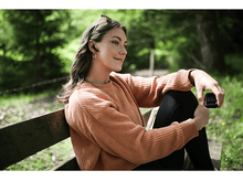 Charger l&#39;image dans la galerie, Une femme portant des écouteurs sourit, assise sur un banc.
