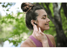 Charger l&#39;image dans la galerie, Une femme avec des écouteurs dans l&#39;oreille, en touchant un. Les arbres sont en arrière-plan.
