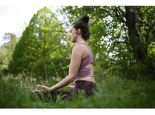 Charger l&#39;image dans la galerie, Femme dans un parc, méditant, portant des écouteurs, entourée d&#39;herbe verte.
