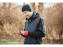 Charger l&#39;image dans la galerie, Un homme en veste et bonnet noirs tient des écouteurs, souriant. Arrière-plan flou.
