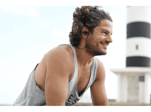 Charger l&#39;image dans la galerie, Homme portant des écouteurs, souriant, avec un phare en arrière-plan.
