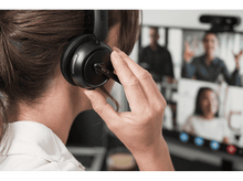 Charger l&#39;image dans la galerie, Femme portant un casque lors d&#39;un appel vidéo. D&#39;autres personnes sont visibles sur un écran en arrière-plan.
