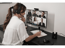 Charger l&#39;image dans la galerie, Une femme en chemise blanche est en appel vidéo, prend des notes et porte un casque.

