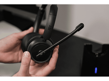 Charger l&#39;image dans la galerie, Mains tenant un casque noir avec un microphone. Une base de chargement est visible en arrière-plan.
