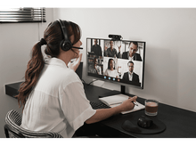Charger l&#39;image dans la galerie, Femme en appel vidéo avec des collègues sur un écran d&#39;ordinateur. Elle porte un casque et gesticule.
