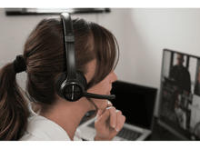 Charger l&#39;image dans la galerie, Femme avec casque et micro, participant à un appel vidéo sur un ordinateur portable. Participants flous en arrière-plan.
