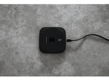 Charger l'image dans la galerie, Un chargeur sans fil carré noir avec le logo ISY, branché sur un câble noir sur une surface en béton grise.
