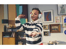 Charger l&#39;image dans la galerie, Un jeune garçon portant une chemise rayée et un casque, gesticulant avec les mains, entouré d&#39;art et de meubles.
