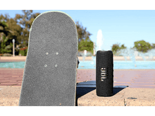 Charger l&#39;image dans la galerie, Enceinte JBL noire à côté d&#39;un skateboard. En arrière-plan, une fontaine et des arbres.
