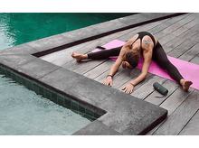 Charger l&#39;image dans la galerie, Femme faisant du yoga près d&#39;une piscine. Elle est sur un tapis rose, jambes écartées, touchant ses pieds.
