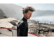 Charger l&#39;image dans la galerie, Un homme en combinaison de plongée tenant une planche de surf sur une plage. La mer est derrière lui.

