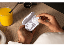 Charger l'image dans la galerie, Personne tenant des écouteurs JBL blancs dans un étui de chargement blanc. Un verre de jus d'orange à proximité.
