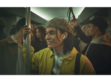 Charger l'image dans la galerie, Homme dans un train portant des écouteurs. Gens en arrière-plan. Portant une veste jaune et une casquette.
