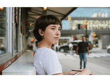 Charger l'image dans la galerie, Une jeune femme avec des écouteurs, regardant ailleurs, assise à une table dans une rue de la ville.
