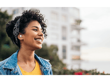 Charger l'image dans la galerie, Femme avec des écouteurs souriant, portant une veste en jean. Fond : vue urbaine.
