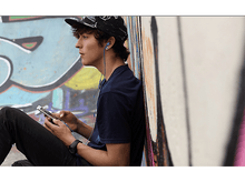 Charger l&#39;image dans la galerie, Un jeune homme avec des écouteurs et un téléphone est assis près d&#39;un mur de graffitis.
