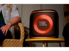 Charger l&#39;image dans la galerie, Un haut-parleur JBL avec un anneau lumineux orange est sur une table jaune. Une personne est à proximité.
