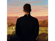 Le dos d'un homme face à un paysage désertique. Il porte un costume noir. Le ciel montre un coucher de soleil.