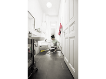 Charger l&#39;image dans la galerie, Une femme nettoie le sol d&#39;une salle de bain avec une machine de nettoyage. La pièce est blanche avec des carreaux sombres.
