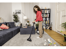 Charger l&#39;image dans la galerie, Femme passant l&#39;aspirateur sur un tapis dans un salon. Des jouets sont éparpillés sur le sol. Canapé et bibliothèque en arrière-plan.

