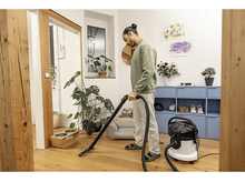 Charger l&#39;image dans la galerie, Un homme passe l&#39;aspirateur sur un sol en bois dans une pièce bien éclairée, avec plantes et meubles.
