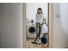 Charger l&#39;image dans la galerie, Un homme passe l&#39;aspirateur dans une pièce à travers une porte. Une machine à laver et des produits de nettoyage sont à l&#39;arrière-plan.
