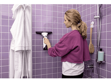 Charger l&#39;image dans la galerie, Femme avec un nettoyeur de vitres essuyant des carreaux violets dans la douche, peignoir pendu.
